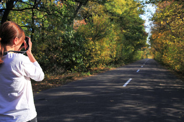 Golden autumn has come. Young girl is taking photography of the picturesque autumn road during the travelling. Colorful landscape of the country road, fallen leaves and yellow trees tunnel