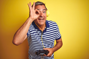 Middle age man playing video game using headphones over isolated yellow background with happy face smiling doing ok sign with hand on eye looking through fingers