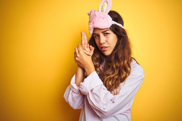 Young woman wearing pajama and sleep mask standing over yellow isolated background Holding symbolic gun with hand gesture, playing killing shooting weapons, angry face