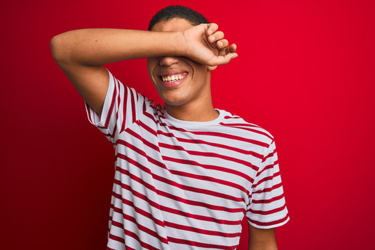 Young handsome arab man wearing striped t-shirt over isolated red background covering eyes with arm smiling cheerful and funny. Blind concept.