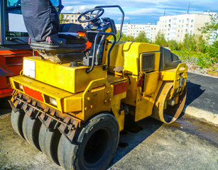 A yellow small self-propelled asphalt roller stands in an open-air car park on a weekend.