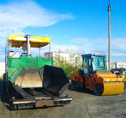 Road machinery - asphalt paver machine and asphalt roller are parked in a parking lot awaiting workers.