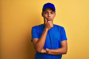 Young handsome arab delivery man standing over isolated yellow background looking confident at the camera with smile with crossed arms and hand raised on chin. Thinking positive.