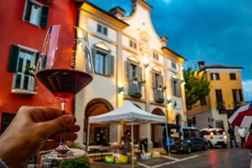 hand holding a glass of red wine in the town of Neive, Italy, one of the main villages of the Langhe hills, famous district of Barolo and Barbaresco wine. - obrazy, fototapety, plakaty