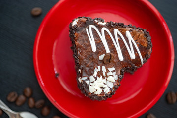 A delicious heart-shaped cake sprinkled with pieces of chocolate lies on a red plate. Photographed close-up on a wooden background.