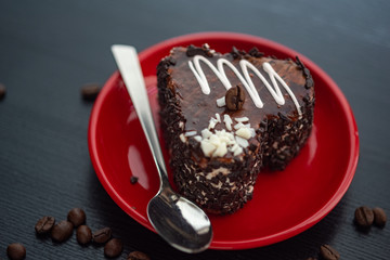 A delicious heart-shaped cake sprinkled with pieces of chocolate lies on a red plate. Photographed close-up on a wooden background.