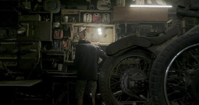 Authentic motorcycle master works as a grinder in an old workshop against a background of vintage motorcycles.