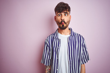 Young man with tattoo wearing striped shirt standing over isolated pink background making fish face with lips, crazy and comical gesture. Funny expression.