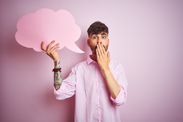 Young man with tattoo holding cloud speech bubble standing over isolated pink background cover mouth with hand shocked with shame for mistake, expression of fear, scared in silence, secret concept