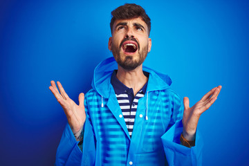 Young handsome man wearing rain coat standing over isolated blue background crazy and mad shouting and yelling with aggressive expression and arms raised. Frustration concept.