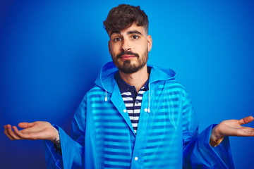 Young handsome man wearing rain coat standing over isolated blue background clueless and confused expression with arms and hands raised. Doubt concept.