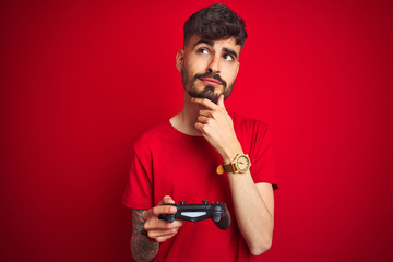 Young gamer man with tattoo playing video game standing over isolated red background serious face...