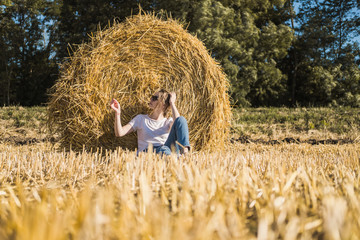 Naklejka na ściany i meble Young woman with brown hair in white T-shirt and blue jeans poses by haystack on a field after harvest. Warm sunset lighting.