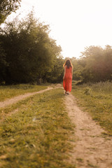 Beautiful young girl with dark curly hair in bright orange dress walks into the distance through forest or field at sunset. Romantic mood. Loneliness.