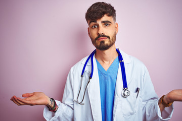 Young doctor man with tattoo wearing stethocope standing over isolated pink background clueless and confused expression with arms and hands raised. Doubt concept.