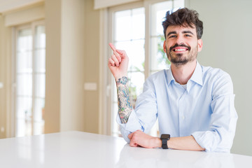 Young businesss man sitting on white table with a big smile on face, pointing with hand and finger to the side looking at the camera.