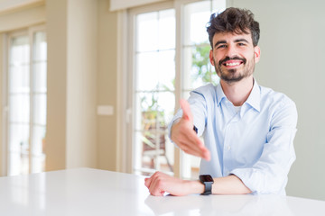 Young businesss man sitting on white table smiling friendly offering handshake as greeting and welcoming. Successful business.