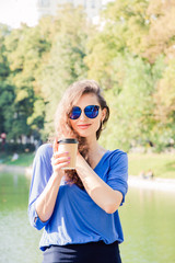 Beautiful brunette woman posing near the pond in park with a Cup of coffee in summertime. Portrait of a young woman