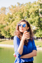 Beautiful brunette woman posing near the pond in park with a Cup of coffee in summertime. Portrait of a young woman