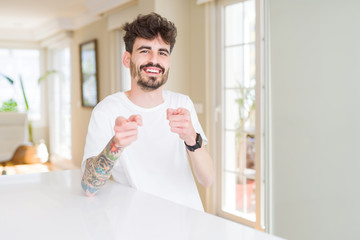 Young man wearing casual t-shirt sitting on white table pointing fingers to camera with happy and funny face. Good energy and vibes.