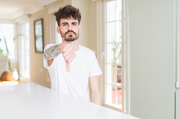 Young man wearing casual t-shirt sitting on white table looking unhappy and angry showing rejection and negative with thumbs down gesture. Bad expression.