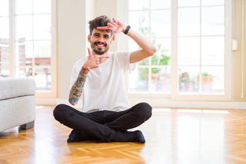 Young man working sitting casual on the floor at home smiling making frame with hands and fingers with happy face. Creativity and photography concept.