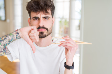 Young man eating asian sushi using chopsticks with angry face, negative sign showing dislike with...