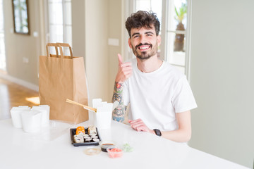 Young man eating asian sushi from home delivery doing happy thumbs up gesture with hand. Approving expression looking at the camera with showing success.