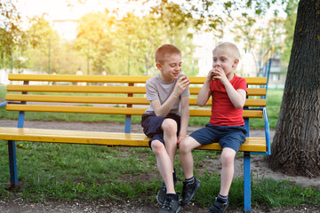 Two boys have a snack on a bench in the park and chat. Summer day