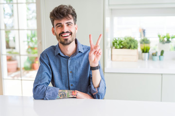 Young man wearing casual shirt sitting on white table smiling with happy face winking at the camera doing victory sign. Number two.