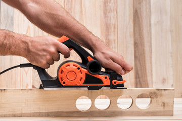 Electric planer in male hand. Processing of the workpiece on light brown wooden table. Side view