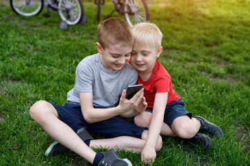 Naklejka na ściany i meble Two boys with a smartphone in their hands are sitting on the grass. Bicycles in the background