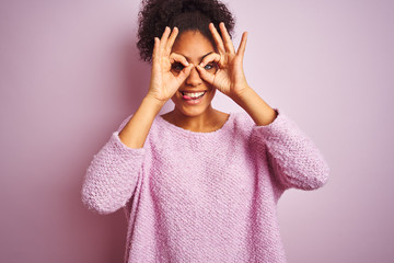 Young african american woman wearing winter sweater standing over isolated pink background doing ok gesture like binoculars sticking tongue out, eyes looking through fingers. Crazy expression.