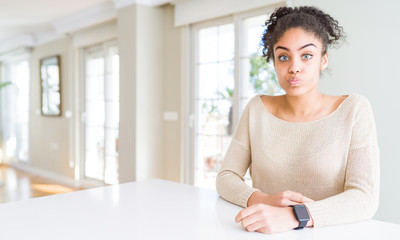 Obraz premium Beautiful young african american woman with afro hair sitting on table at home puffing cheeks with funny face. Mouth inflated with air, crazy expression.