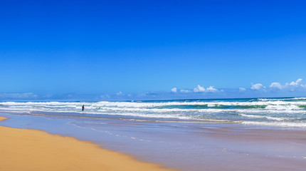 Obraz premium Beautiful wide panoramic view of the ocean waves rolling towards tropical sandy beach and a silhouette of a man standing in the water. Gold Coast, Queensland, Australia. 
