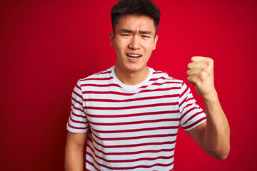 Young asian chinese man wearing striped t-shirt standing over isolated red background angry and mad raising fist frustrated and furious while shouting with anger. Rage and aggressive concept.