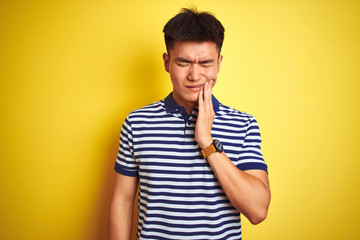 Young asian chinese man wearing striped polo standing over isolated yellow background touching...