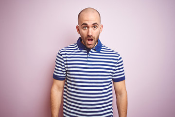 Young bald man with beard wearing casual striped blue t-shirt over pink isolated background afraid and shocked with surprise expression, fear and excited face.