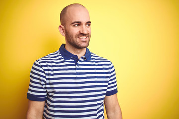 Young bald man with beard wearing casual striped blue t-shirt over yellow isolated background looking away to side with smile on face, natural expression. Laughing confident.