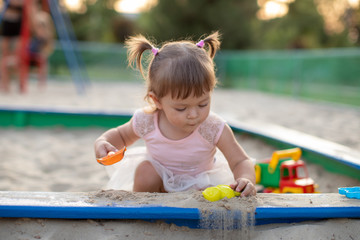 Cute toddler girl playing in sand on outdoor playground. Beautiful baby in red trousers having fun on sunny warm summer day. Child with colorful sand toys. Healthy active baby outdoors plays games
