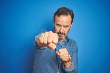 Handsome middle age senior man with grey hair over isolated blue background Punching fist to fight,...
