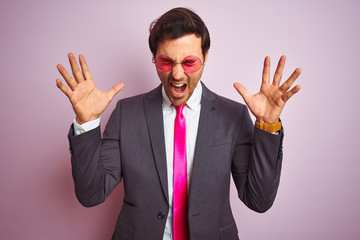 Young handsome businessman wearing suit and sunglasses over isolated pink background celebrating mad and crazy for success with arms raised and closed eyes screaming excited. Winner concept
