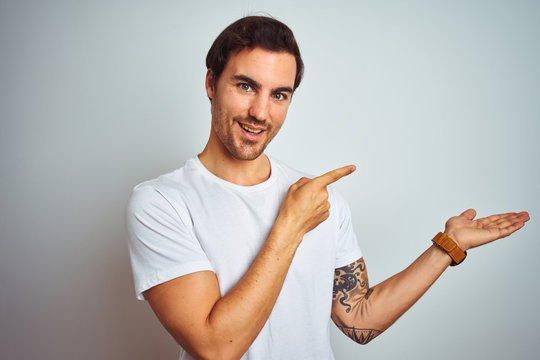 Young handsome man with tattoo wearing casual t-shirt over isolated white background amazed and smiling to the camera while presenting with hand and pointing with finger.