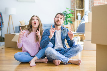 Young beautiful couple moving to a new house sitting on the floor amazed and surprised looking up and pointing with fingers and raised arms.