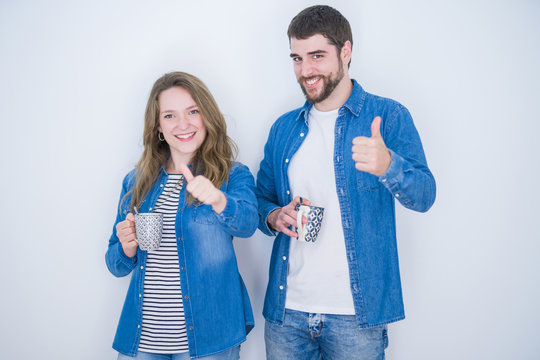 Young beautiful couple drinking a cup of coffee over white isolated background happy with big smile doing ok sign, thumb up with fingers, excellent sign