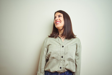 Young beautiful brunette woman wearing green shirt over isolated background looking away to side with smile on face, natural expression. Laughing confident.