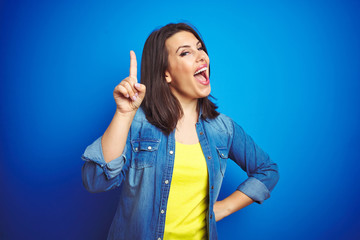 Young beautiful brunette woman wearing casual blue denim jacket over blue isolated background pointing finger up with successful idea. Exited and happy. Number one.