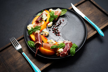 Gourmet salad with blue cheese, peaches, jamon (ham) and basil leaves on a black plate. Dark background