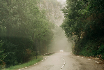 White car silhouette driving fast on mountain road in the Vercors Massif on a misty day with fog and tall mountain in background
