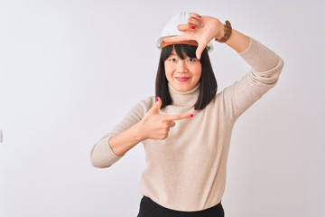 Young beautiful Chinese architect woman wearing helmet over isolated white background smiling making frame with hands and fingers with happy face. Creativity and photography concept.